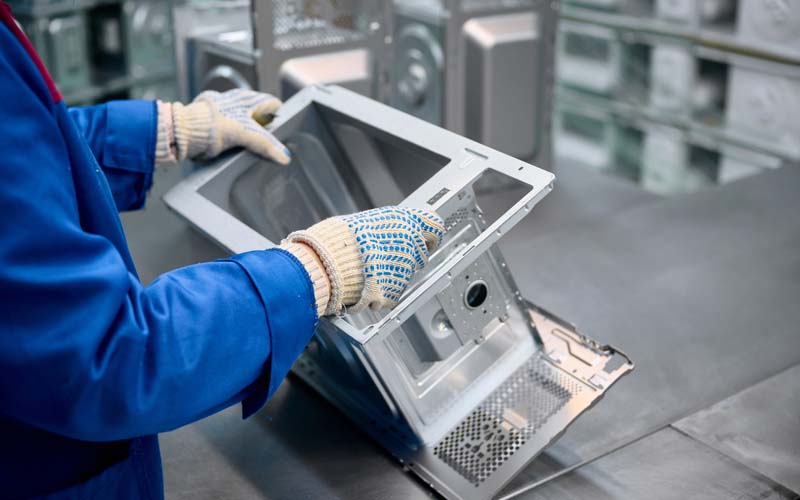 Worker assembles microwave oven casing during manufacturing process in a factory setting. A factory worker carefully assembles the casing of a microwave oven, ensuring precision during the production process. The environment is busy and industrial.